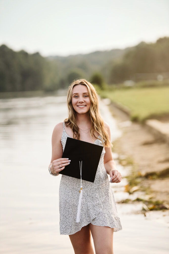 senior girl hold graduation cap at the lake for senior photo session