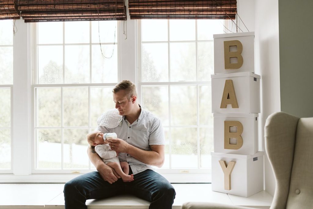 A newborn lifestyle image of a father sitting on a window seat feeding his newborn baby boy. Photo by Pittsburgh Newborn Lifestyle Photographer, Laura Mares Photography.