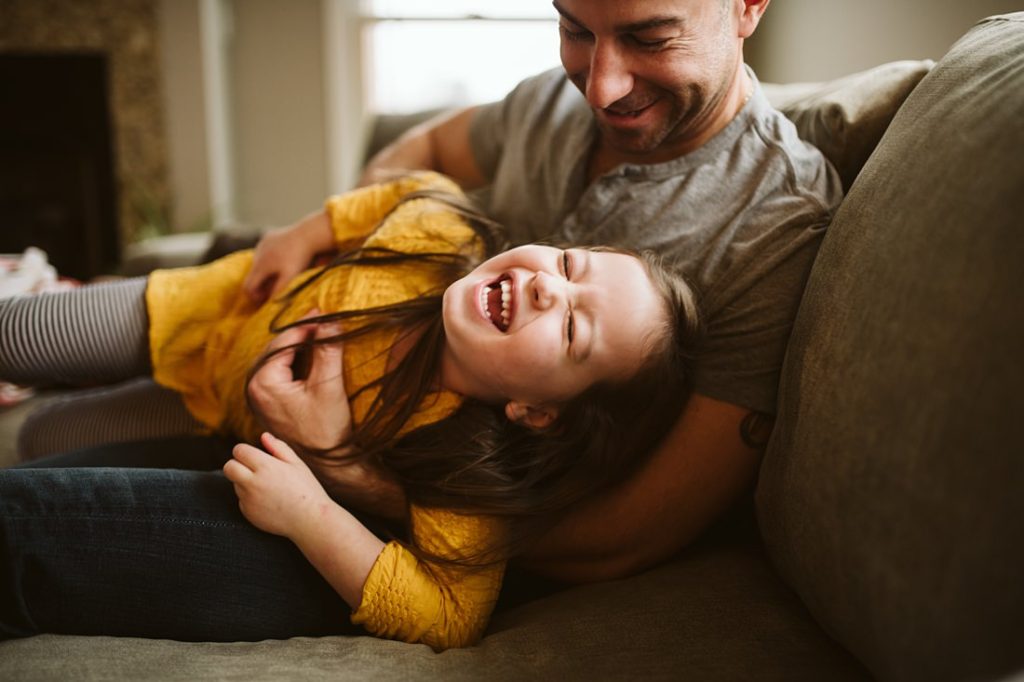 A lifestyle image of a father sitting on the sofa tickling his daughter. Photo by Pittsburgh Lifestyle Photographer, Laura Mares Photography.