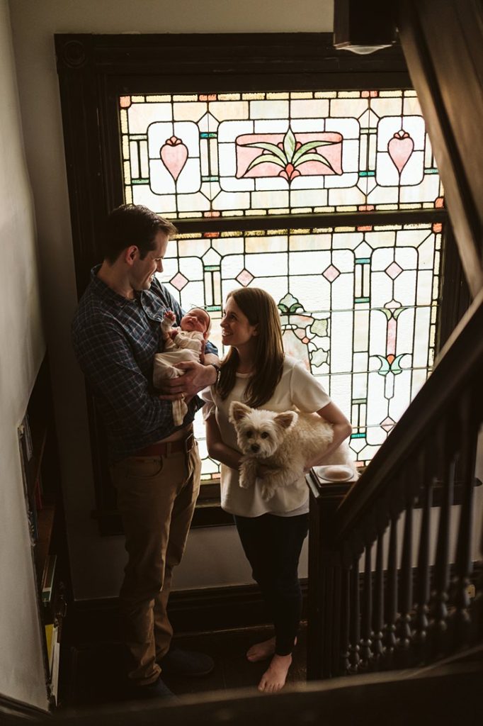 A newborn lifestyle image of a family standing near a stained glass window in the stairway holding their newborn baby. Photo by Pittsburgh Newborn Lifestyle Photographer, Laura Mares Photography.