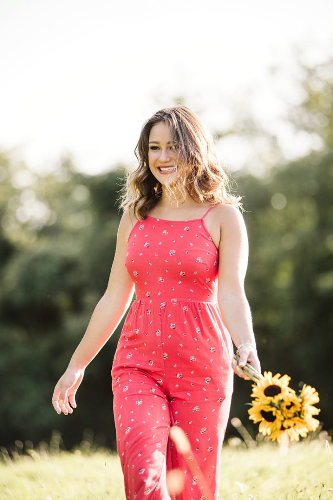 Portrait of a high school senior girl holding sunflowers walking on a field at sunset. Photo by Laura Mares Photography, Pittsburgh Senior Photographer.