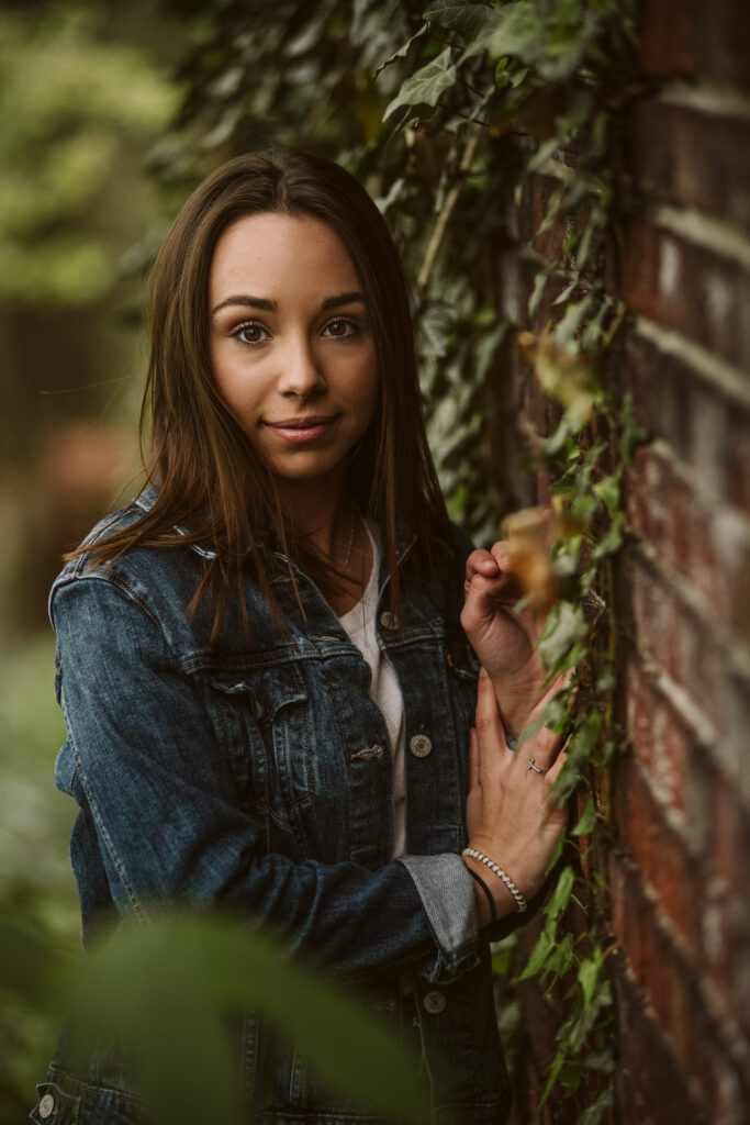Portrait of a senior girl wearing a blue jean jacket standing near a brick wall covered in greenery