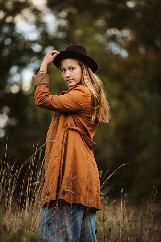 a high school senior girl wearing a trench coat and cowgirl hat standing in a beautiful field at sunset