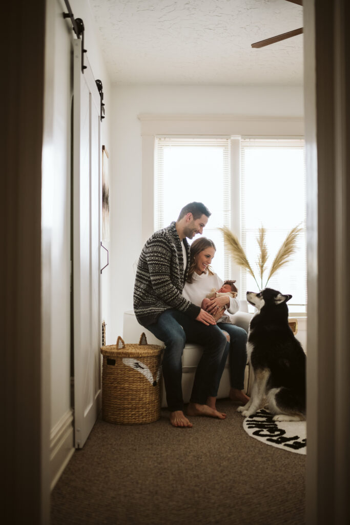 beautiful family holding newborn baby in modern boho nursery with their siberian husky looking at their baby
