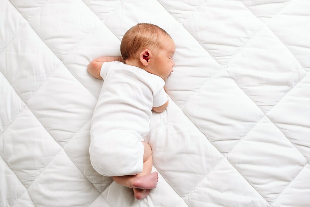newborn portrait of a baby wearing a white onesie sleeping on a white bed