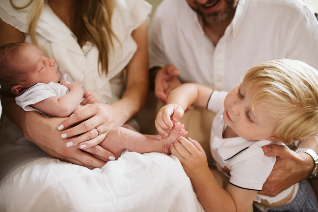 lifestyle portrait of a toddler looking at newborn brother's toes while mom and dad hold both children