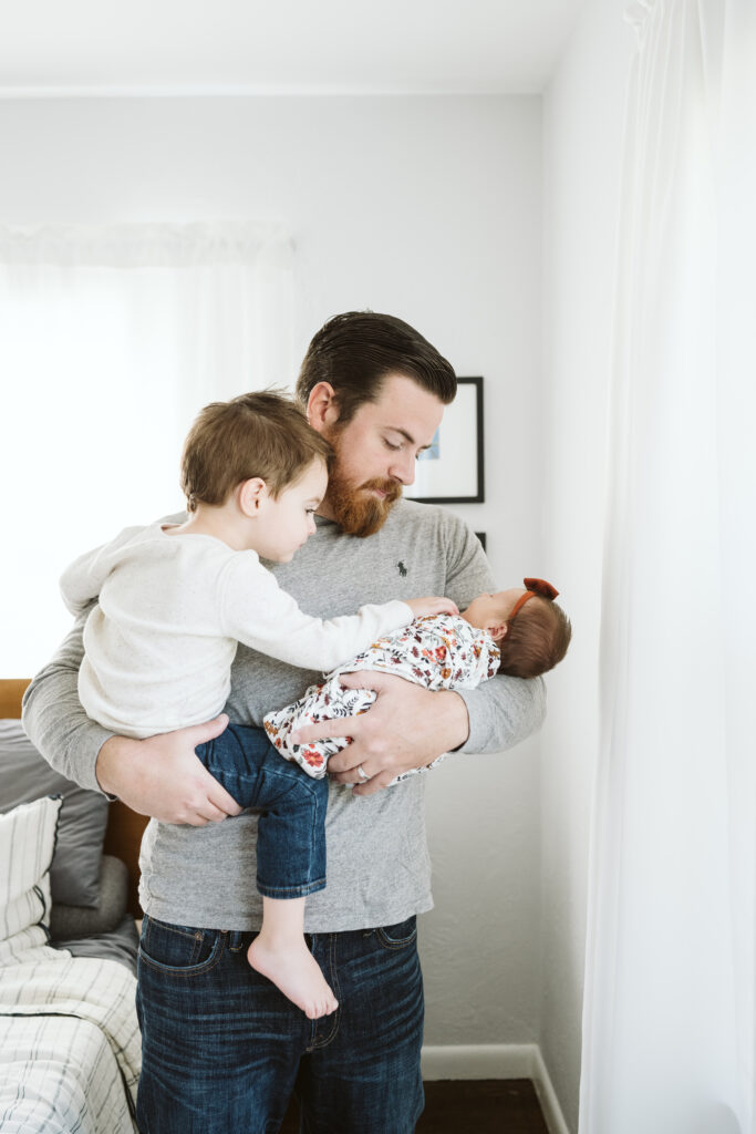 Pittsburgh dad holding newborn baby and big brother near window