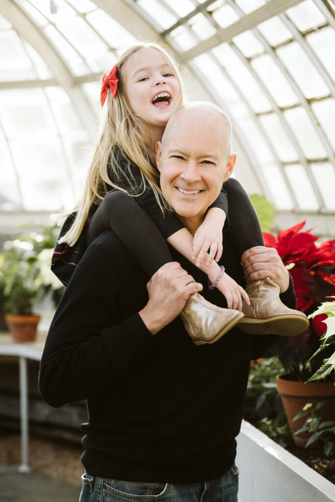 father holding laughing daughter on his shoulders during portrait session in green house