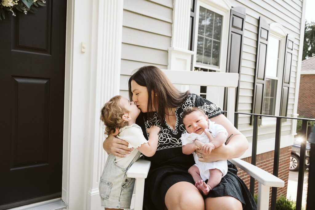 newborn baby held by mom receiving a kiss from her toddler